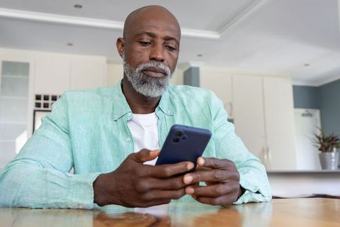 Senior african american man using smartphone in modern kitchen looking focused and thoughtful