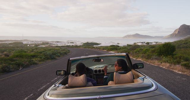 Rear view of diverse couple driving on sunny day in convertible car raising their arms in the air. summer road trip on a country highway by the coast.