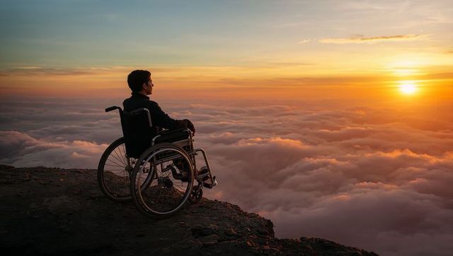 Wheelchair user watching sunset above sea of clouds on mountain summit contemplating horizon