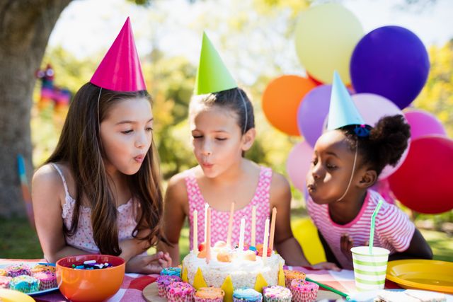 Three girls wearing party hats blowing out candles on a birthday cake at an outdoor party. The scene is festive with colorful balloons and cupcakes on the table. Ideal for use in materials related to children's parties, celebrations, friendship, and outdoor activities.