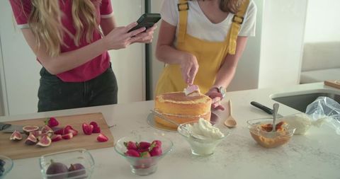 Female friends decorating layered cake in bright home kitchen using smartphone for recipe