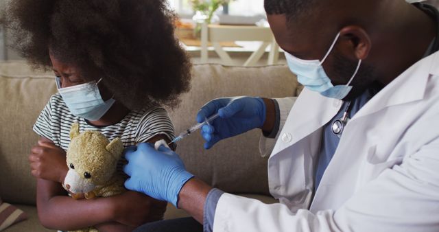 Child receiving vaccine from healthcare professional at home, wearing masks. Child looking upset, hugging teddy bear for comfort. Suitable for promoting pediatric healthcare, vaccination campaigns, home visits, children's medical care, or public health awareness.
