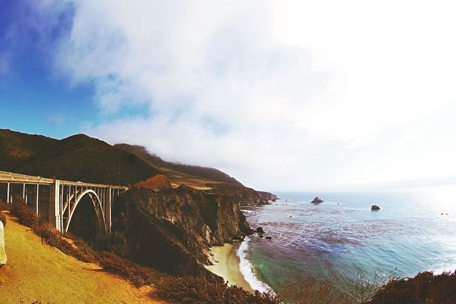 This picturesque scene features a scenic bridge stretching across a stunning coastal cliff. The image captures the rugged beauty of the coastline with waves hitting the cliffs and a misty horizon in the distance. Ideal for travel websites, advertisements for road trips, nature blogs, and promotional materials for coastal tours.