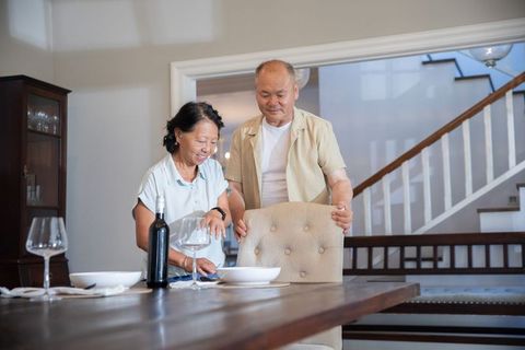 Senior asian couple setting cozy dining table with wine and tufted chair in home