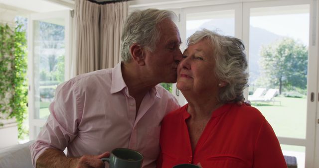 Elderly couple enjoys a tender moment as man kisses woman's cheek while holding coffee mugs, suggesting a morning or daytime scene at home. Bright and airy room with a view of the garden in the background emphasizes a peaceful, loving atmosphere. Perfect for use in campaigns related to senior living, health and wellness, retirement planning, family life, or advertisements aimed at promoting products for older adults.