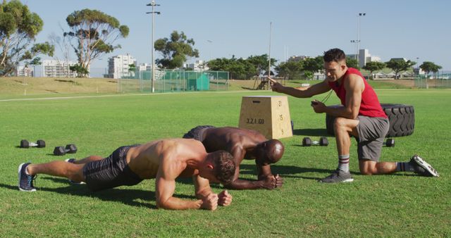 Two men are performing plank exercises on a grassy field while a fitness instructor is coaching and motivating them. This can be used for articles or ads focusing on outdoor fitness training, group workouts, personal training services, and strength training exercises.