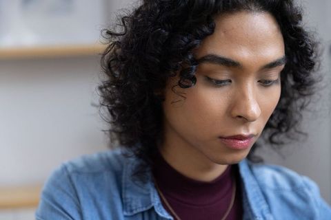 Contemplative non-binary young adult looking downward wearing denim shirt and maroon top