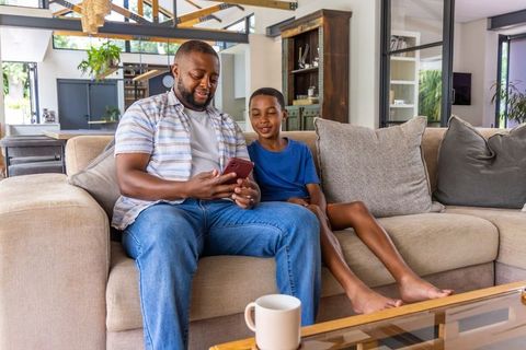 African american father and son sharing smartphone on sofa in modern open-plan living room, natural-