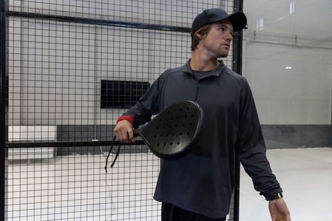Focused young padel player holding racket by mesh fence in indoor court