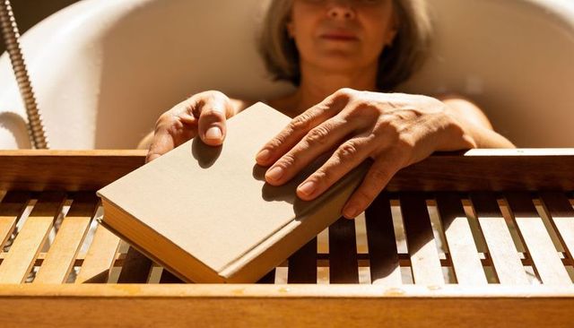 Senior woman relaxing in sunlit bathtub reaching for book on wooden tray