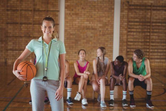 Female coach holding basketball in gym with team sitting in background. Ideal for use in sports training materials, coaching advertisements, team-building promotions, and fitness-related content.