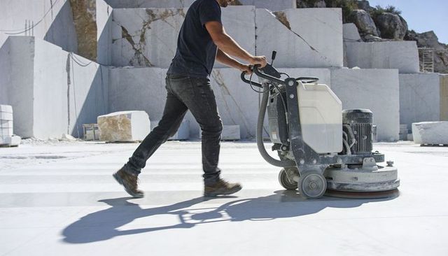 Worker pushing industrial floor grinder polishing white marble surface in outdoor quarry