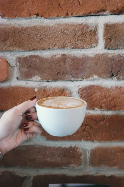 Hand presenting a cappuccino with latte art in a white cup against a rustic brick wall. Perfect for promoting cafes, coffee shops, morning routines, and cozy atmospheres.