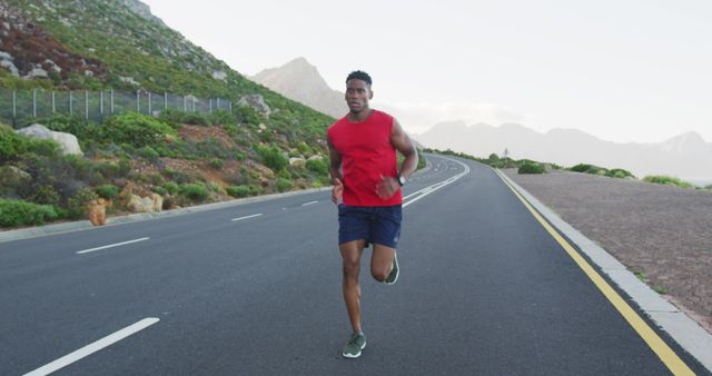 A man is jogging alone on an open country road with mountains in the background. He is wearing a red sleeveless shirt and blue shorts, signifying an active lifestyle. This can be used for content promoting fitness, health, outdoor activities, and summer training routines.