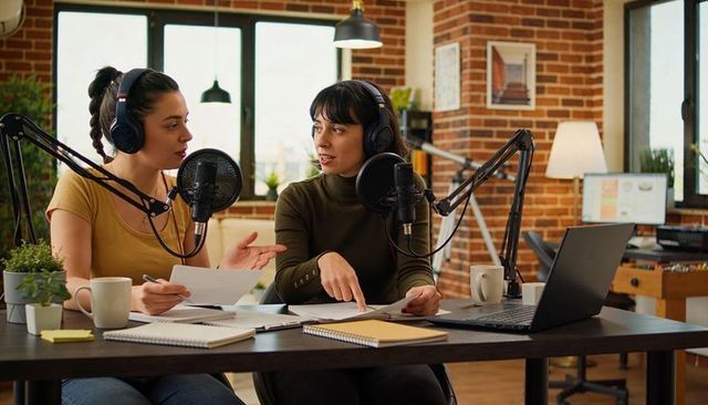 Two cohosts recording podcast in brick loft studio using professional microphones and laptop