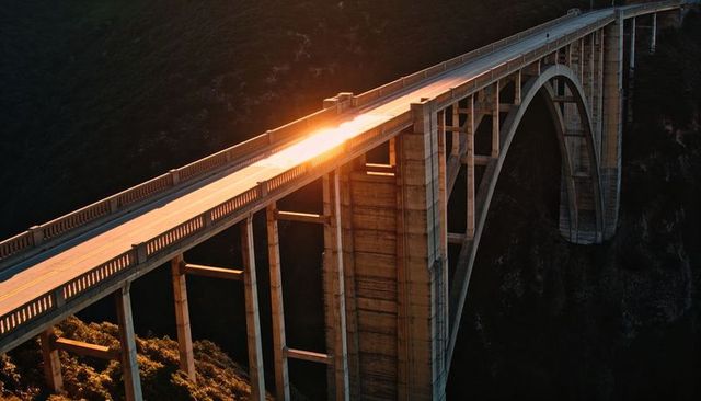 Sunlight reflecting on arched concrete bridge spanning coastal canyon at sunset