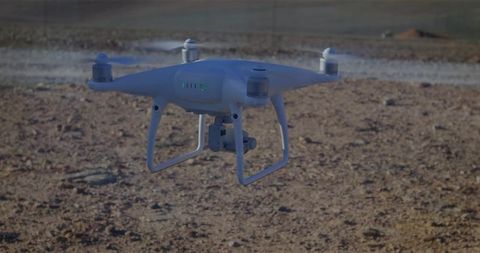 White quadcopter drone hovering over rocky desert field with camera gimbal, cinematic