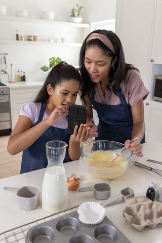 African american mother and daughter baking muffins while checking recipe on smartphone