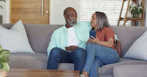Middle-aged african american couple relaxing on sofa sharing smartphone in cozy home