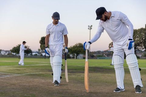 Two diverse cricketers walking off pitch at dusk wearing white kit and protective pads