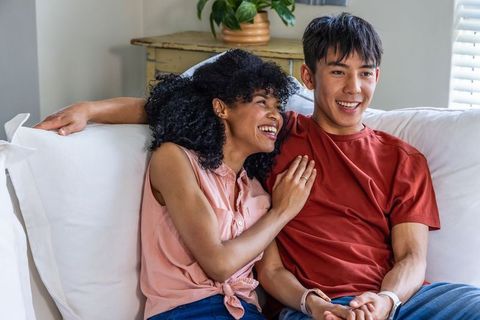 Interracial couple laughing on couch, woman showing engagement ring in cozy living room