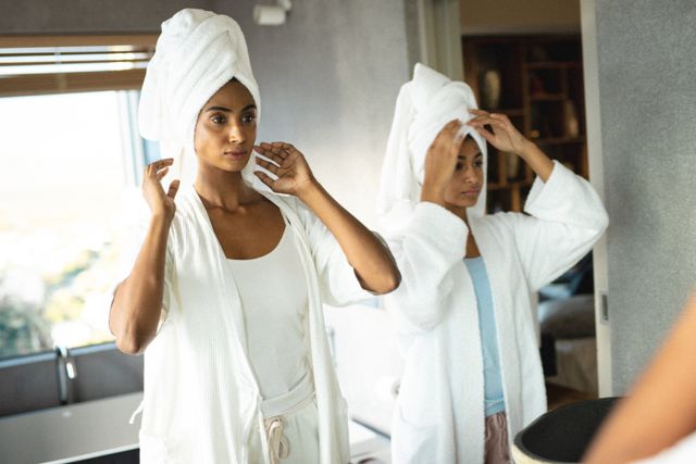 Biracial mother and daughter wearing bathrobes and towels on their heads, standing in front of a mirror in a bathroom. They are engaging in a morning routine, focusing on self-care and bonding. This image can be used for promoting family love, body care products, wellbeing, and self-care routines.