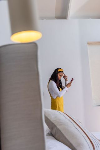 Young woman in yellow overalls checking smartphone while adjusting headband in bedroom