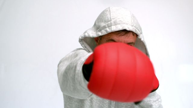 Man wearing hooded sweatshirt and red boxing gloves delivers a strong punch, ideal for use in sports and fitness content. Perfect for illustrating strength, commitment, and active training in gym-related advertising or motivational materials.