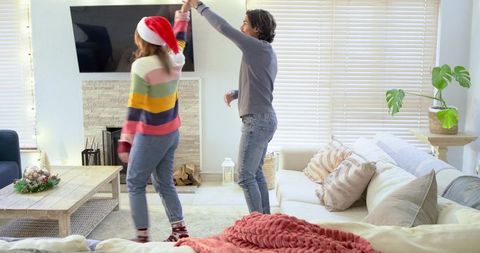 Couple dancing in cozy living room wearing santa hat and colorful sweater holiday romance