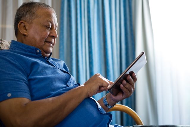 Senior man sitting on sofa using digital tablet in nursing home. Ideal for illustrating technology use among elderly, modern healthcare facilities, and senior lifestyle. Suitable for articles on aging, retirement, and digital inclusion.