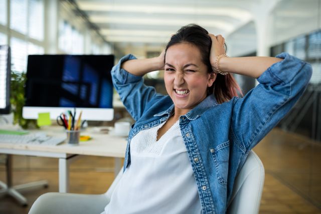 Young female business executive expressing frustration while sitting at her desk in a modern office. Ideal for use in articles about workplace stress, business challenges, and professional environments.