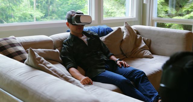 Man is relaxing on comfortable sofa wearing virtual reality headset. Use for themes related to home entertainment, VR technology, relaxation, and modern living.