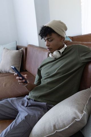 African young man relaxing on sofa scrolling phone wearing beanie and white headphones