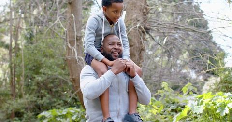 African american father carrying son on shoulders walking through forest trail smiling