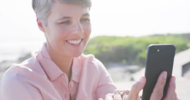 Caucasian woman smiling while using a smartphone during a coastal holiday on a sunny day. Great for illustrating concepts of technology in leisure, retirement activities, and enjoying free time by the sea. Useful for travel adverts, wellness promotions, and lifestyle blogs focusing on mature individuals and digital trends.