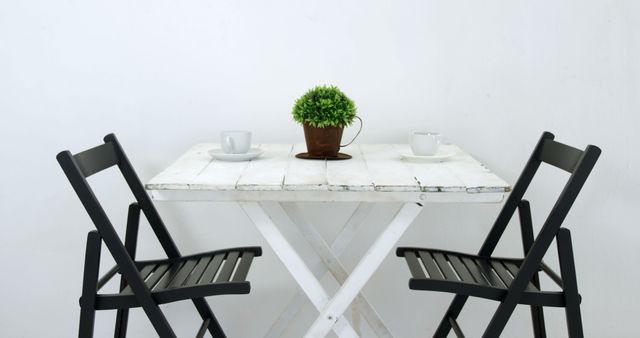 A minimalist dining setup features a white marble table with two black chairs, a small potted plant, and two cups. The clean and simple design creates a serene and inviting space for a meal or conversation.