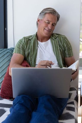 Mature man working from home on laptop writing notes on daybed