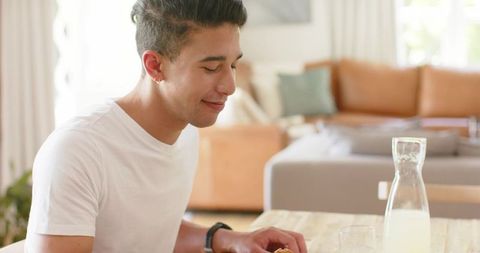 Young man enjoying relaxed breakfast at sunlit dining table with pastry and milk
