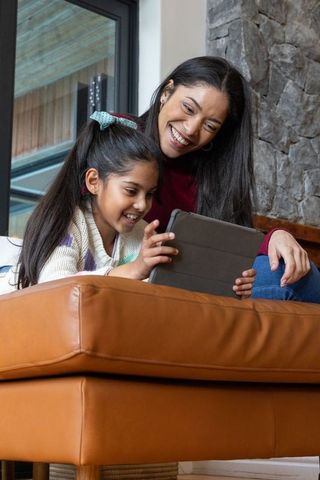 Indian mother and daughter sharing tablet, smiling on leather ottoman in cozy living room