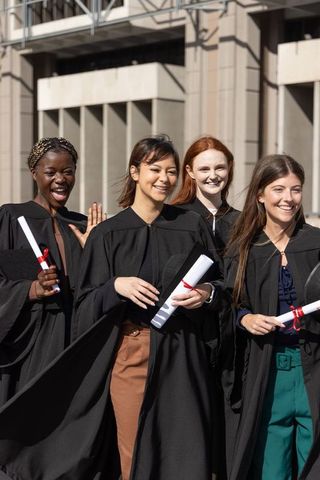Multicultural female graduates celebrating campus plaza in black gowns holding diplomas