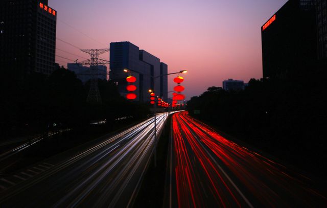 High-resolution image showing an urban highway at dusk with streaking light trails from fast-moving vehicles and streetlights illuminating the scene. High-rise buildings and red lanterns provide an interesting contrast against the night skyline. Ideal for use in articles related to urban life, transportation systems, night photography, or city development. Great for backgrounds, social media banners, and website headers emphasizing modern city infrastructure and night time vibrancy.