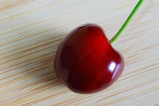 This image features a close-up view of a single, fresh cherry with its stem against a wooden backdrop. The vibrant red color emphasizes its ripeness and juicy texture, perfect for use in advertisements relating to healthy eating, organic produce, nutrition, or summer fruit promotions.