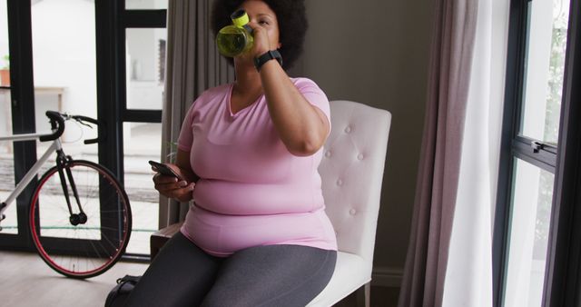 A woman sits on a chair indoors, drinking water from a bottle and looking at her phone. She is wearing casual workout clothes and there is a bicycle in the background. This image can be used for content focusing on women's fitness, hydration importance, at-home workouts, or promoting healthy lifestyles.