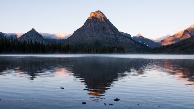 This stunning landscape depicts the Rocky Mountains at sunrise, reflected in a pristine lake with a slight morning mist adding a dreamlike quality. Perfect for use in travel blogs, nature-themed content, desktop wallpapers, or promotional material for outdoor adventure and ecotourism campaigns.