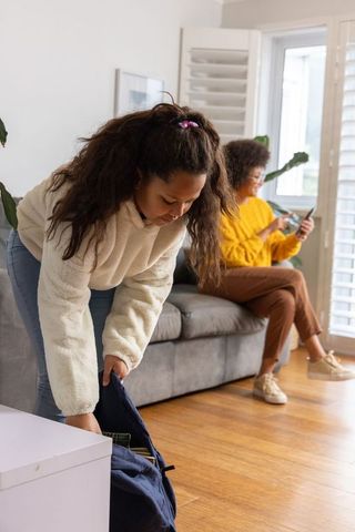African american daughter packing backpack in living room while mother using phone