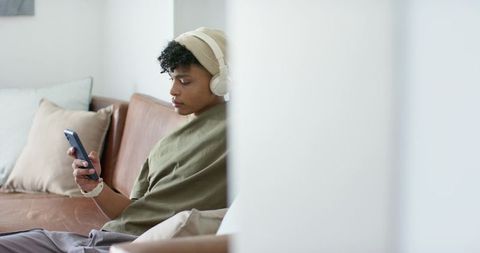 Young african american man sitting on sofa listening to music and checking smartphone