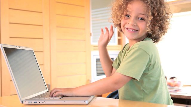 Cheerful boy with curly hair using laptop in a kitchen while waving at camera, ideal for content illustrating children engaging with learning technology, homeschool environments, or playful screenshots in domestic settings.