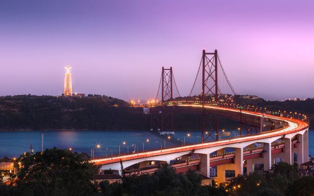 This photo captures the iconic Lisbon suspension bridge with the Christ the King statue in the background, beautifully illuminated at twilight. The long exposure creates a dynamic effect with car light trails. Perfect for use in travel brochures, architecture highlights, urban landscape features, or city guides emphasizing Lisbon's landmarks and nighttime beauty.