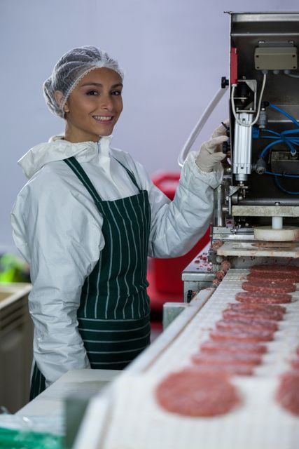 Female butcher wearing safety gear and working in a meat factory processing hamburger patties. Ideal for use in articles about food production, meat industry, food safety, and manufacturing processes.