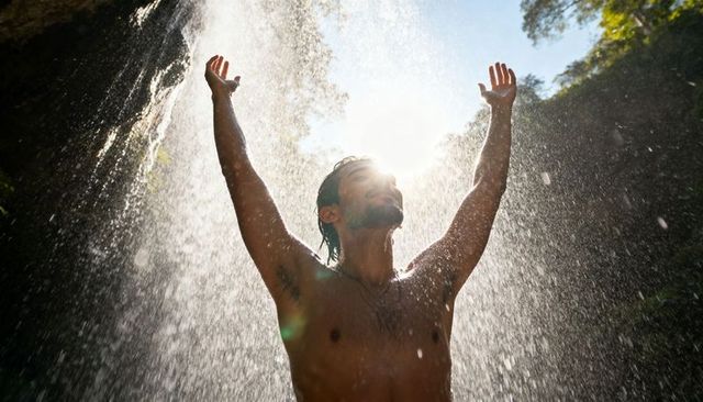 Bare-chested man raising arms under waterfall with sunlight burst